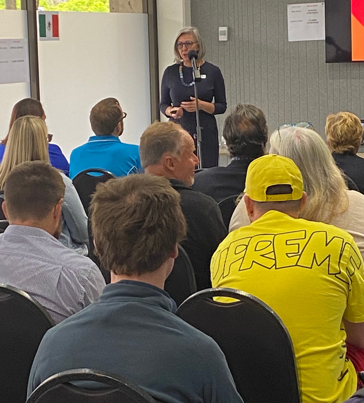Woman speaks at microphone in front of seated audience.